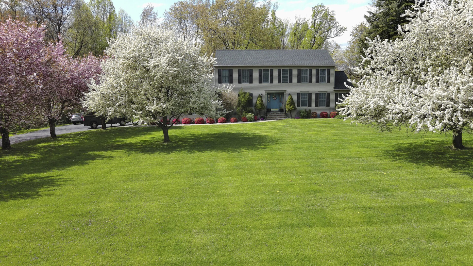 A two-story house with symmetrical windows, surrounded by blooming trees and lush green lawn, under a clear sky. No landmarks or historical buildings.