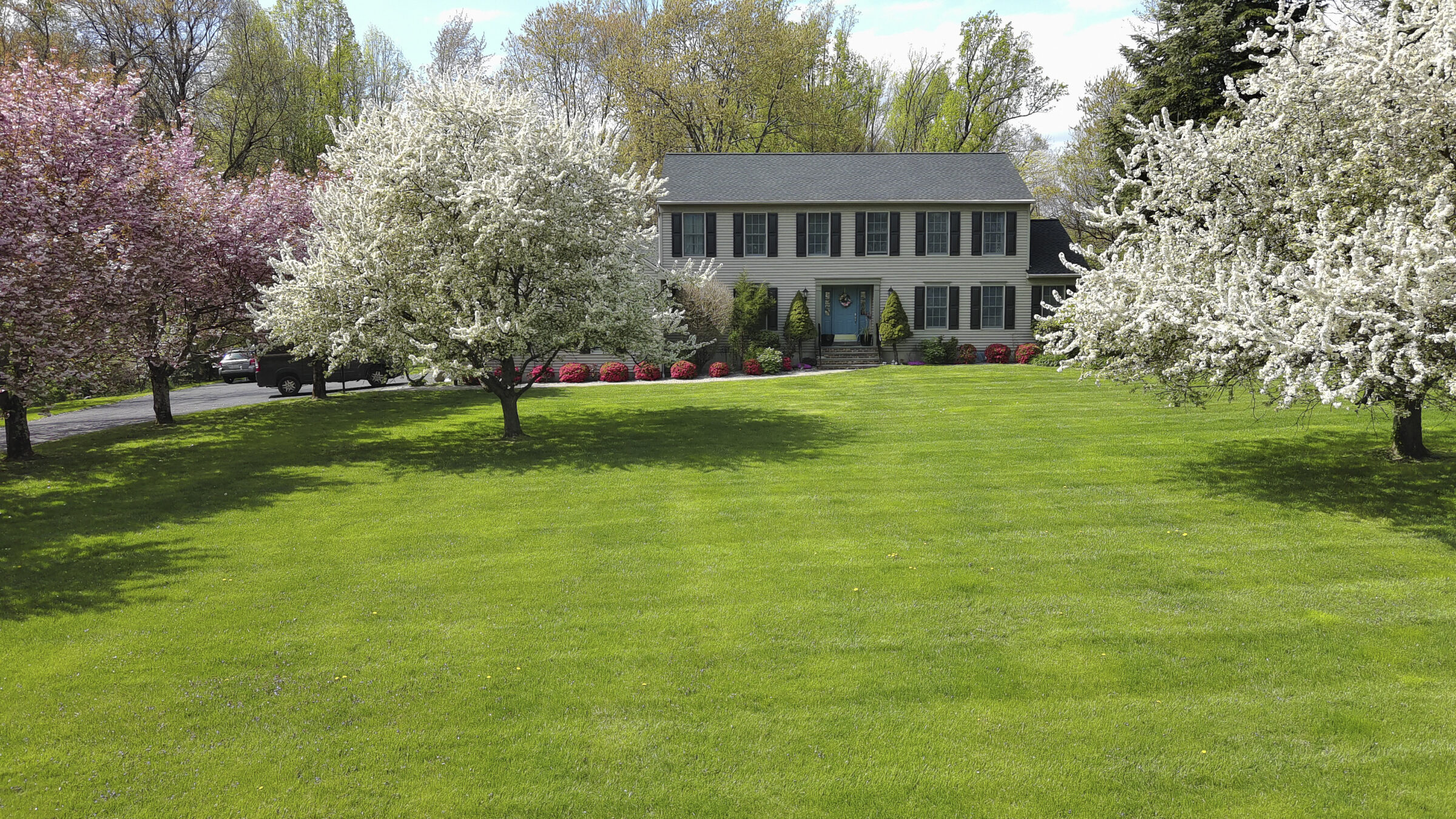 A two-story house with symmetrical windows, surrounded by blooming trees and lush green lawn, under a clear sky. No landmarks or historical buildings.