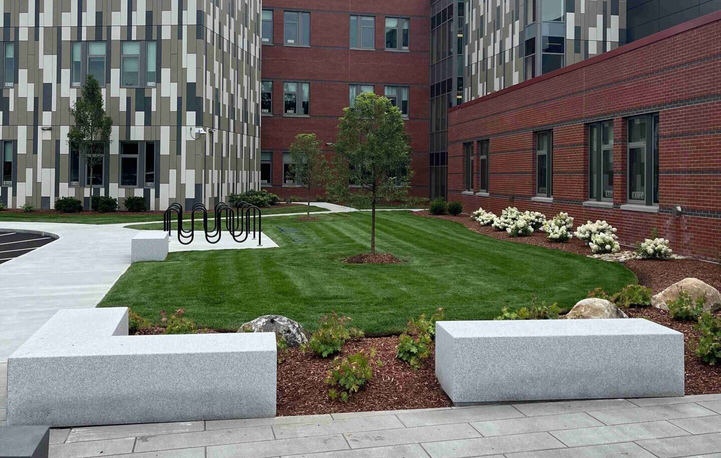 Modern building courtyard with manicured lawn, benches, bike racks, and shrubs. Brick and patterned facades surround the green space, creating a peaceful atmosphere.