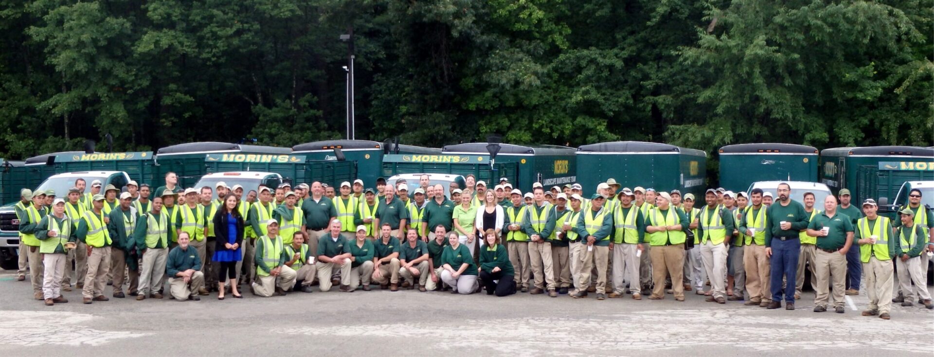 A large group of people in green safety vests stands in front of multiple trucks, with trees in the background.