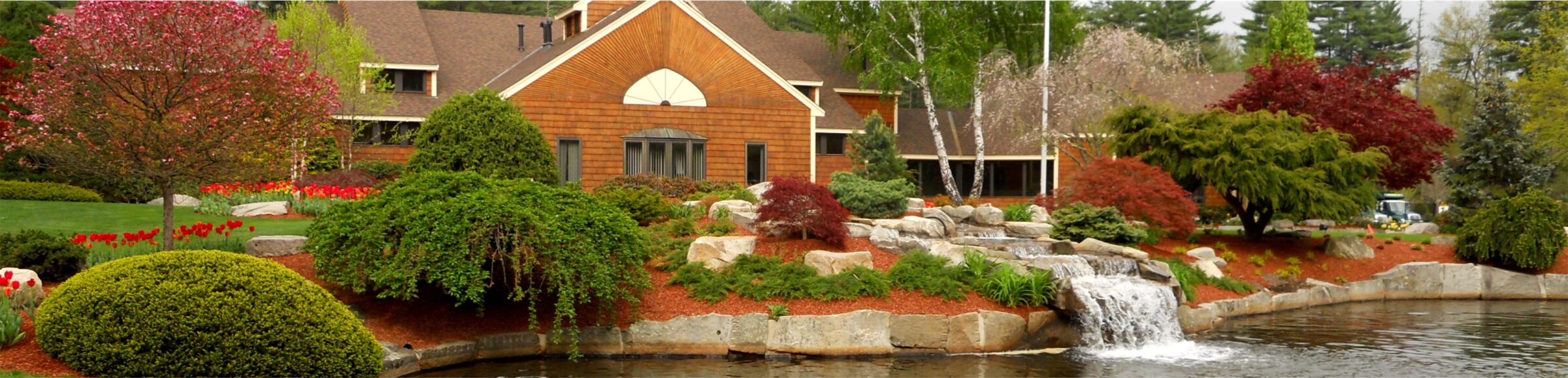 A scenic view of a landscaped garden featuring a wooden building, vibrant greenery, colorful trees, a waterfall, and a calm pond.