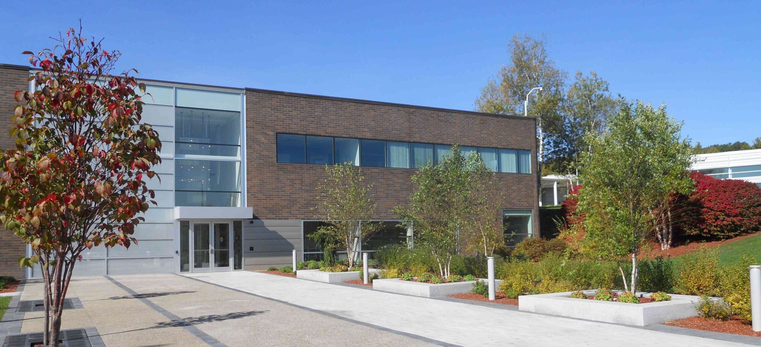 A modern building with large windows, surrounded by landscaped greenery, trees, and a paved walkway under a clear blue sky.