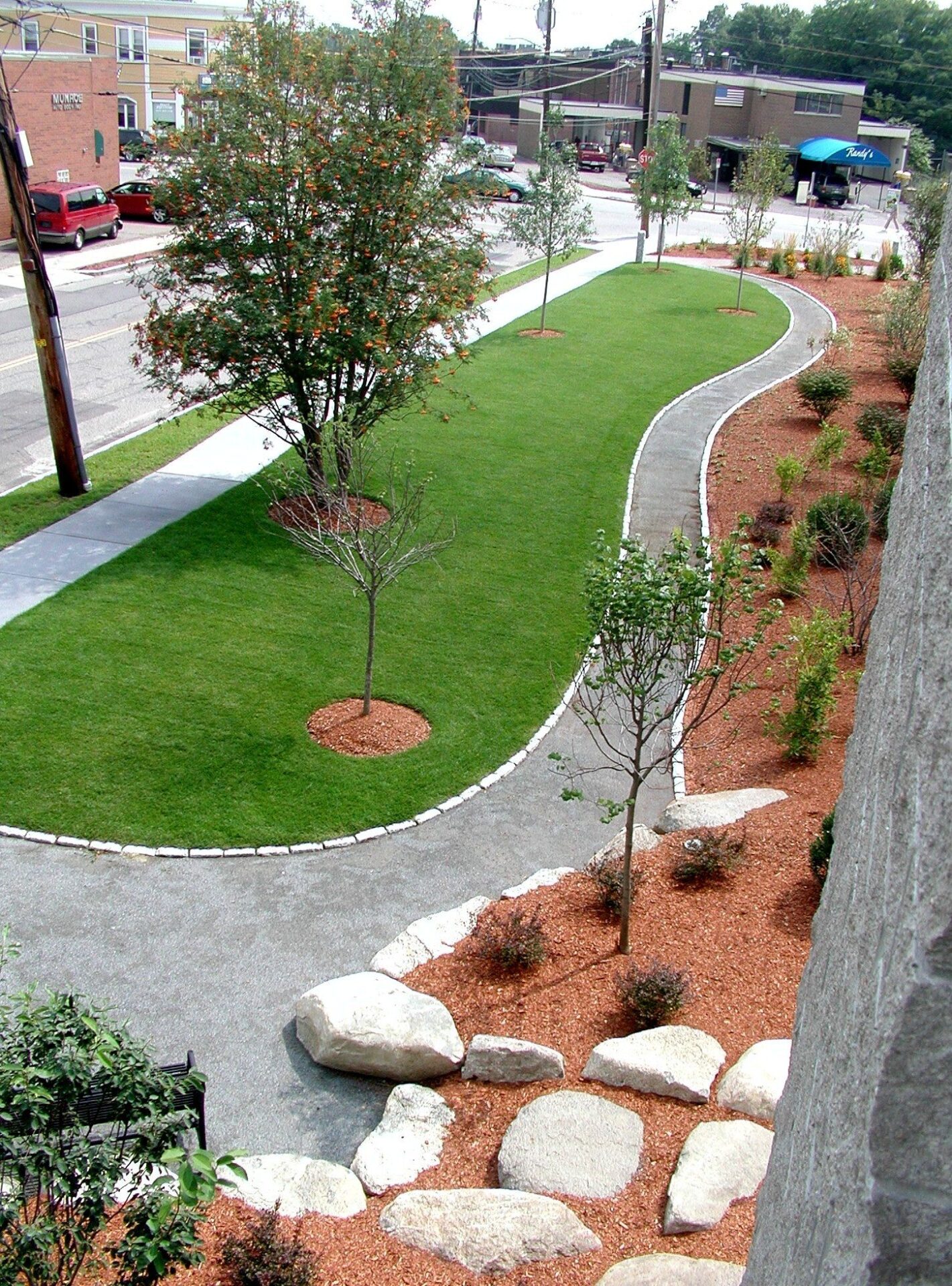 A sidewalk borders a landscaped park with green grass, small trees, and stone accents. Nearby buildings are visible across the street.