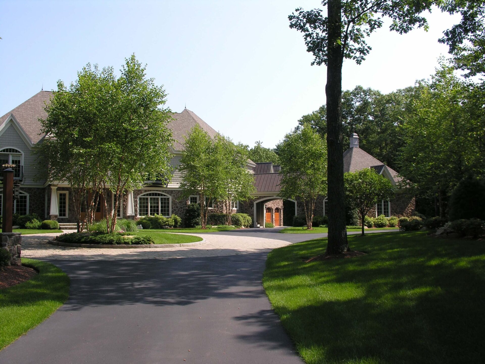 Large estate with circular driveway, surrounded by lush greenery and tall trees. The house has a stone facade and steep, gabled roofs.