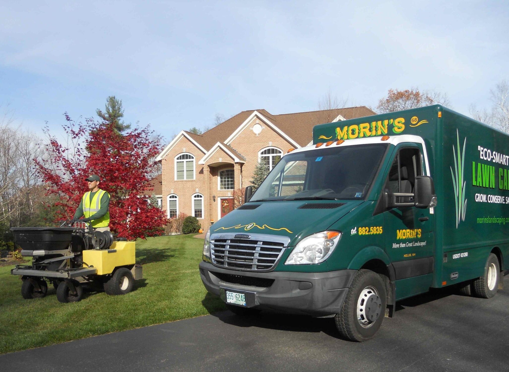 A person operates lawn equipment near a brick house with a red tree nearby. A green van with "Morin's" is parked on the driveway.