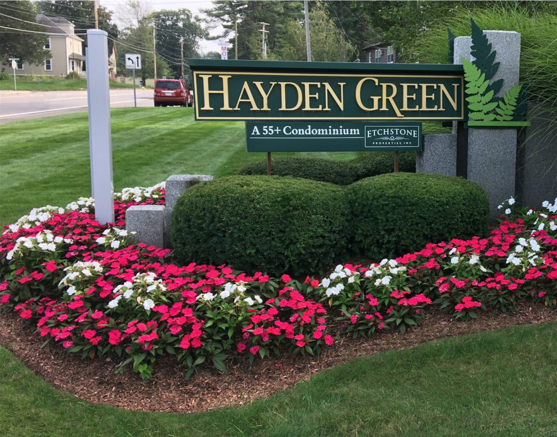 Green lawn with vibrant flower bed surrounds the "Hayden Green" sign for a 55+ condominium. Nearby road and trees visible in the background.
