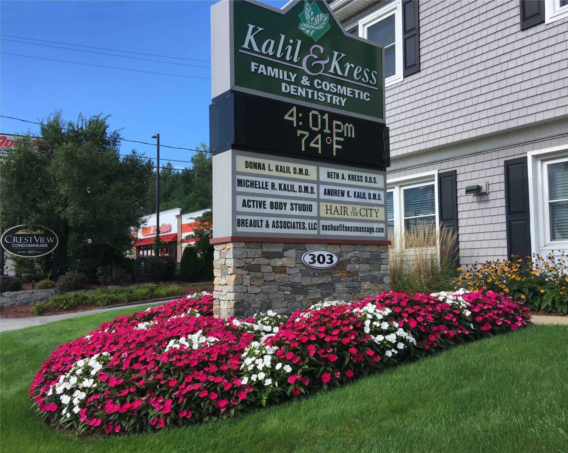 A dentistry office sign surrounded by vibrant pink and white flowers, with a building in the background and a smaller sign nearby.