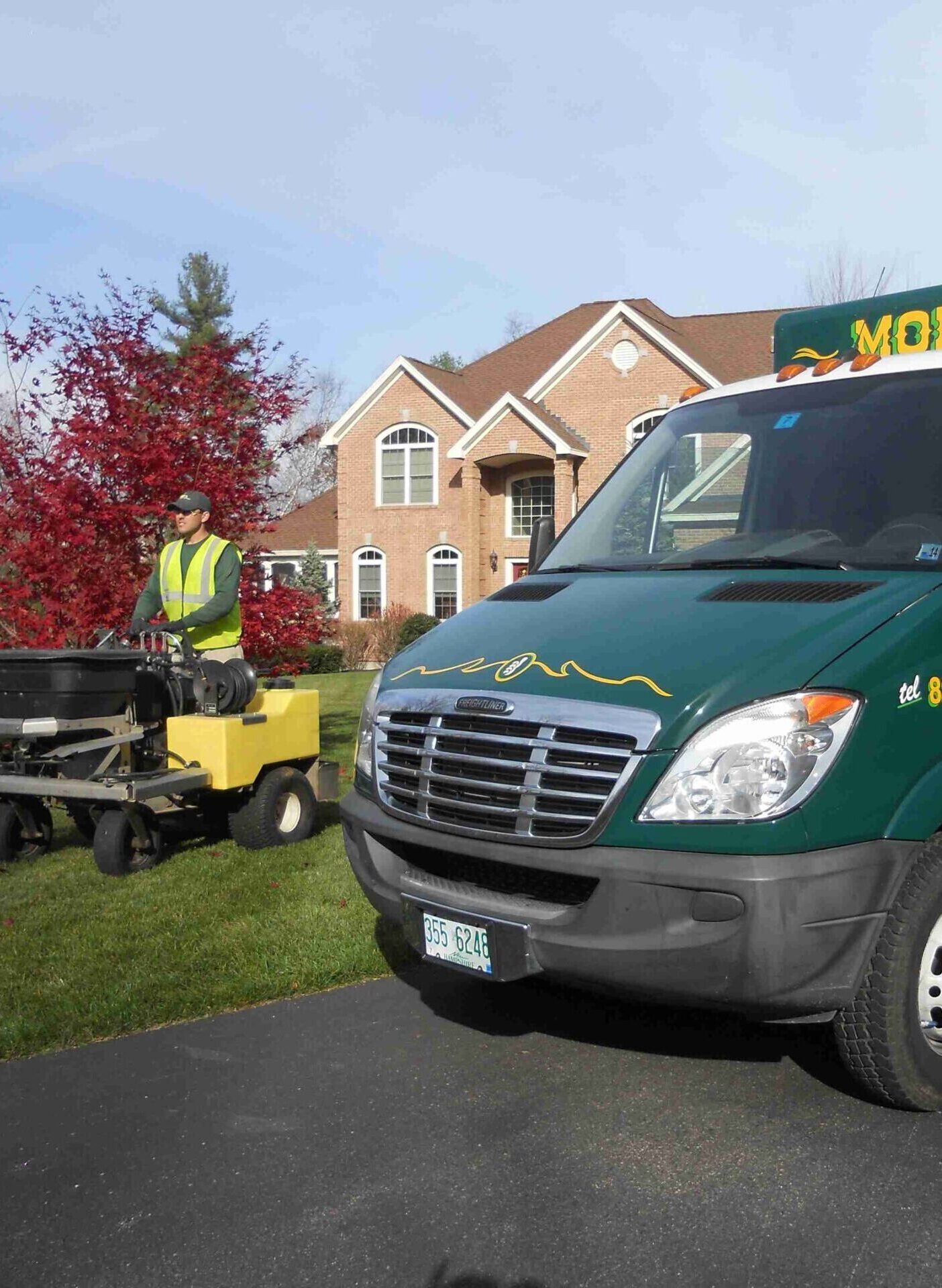 A person operates landscaping equipment on a lawn next to a green service van and a large brick house with red trees in view.