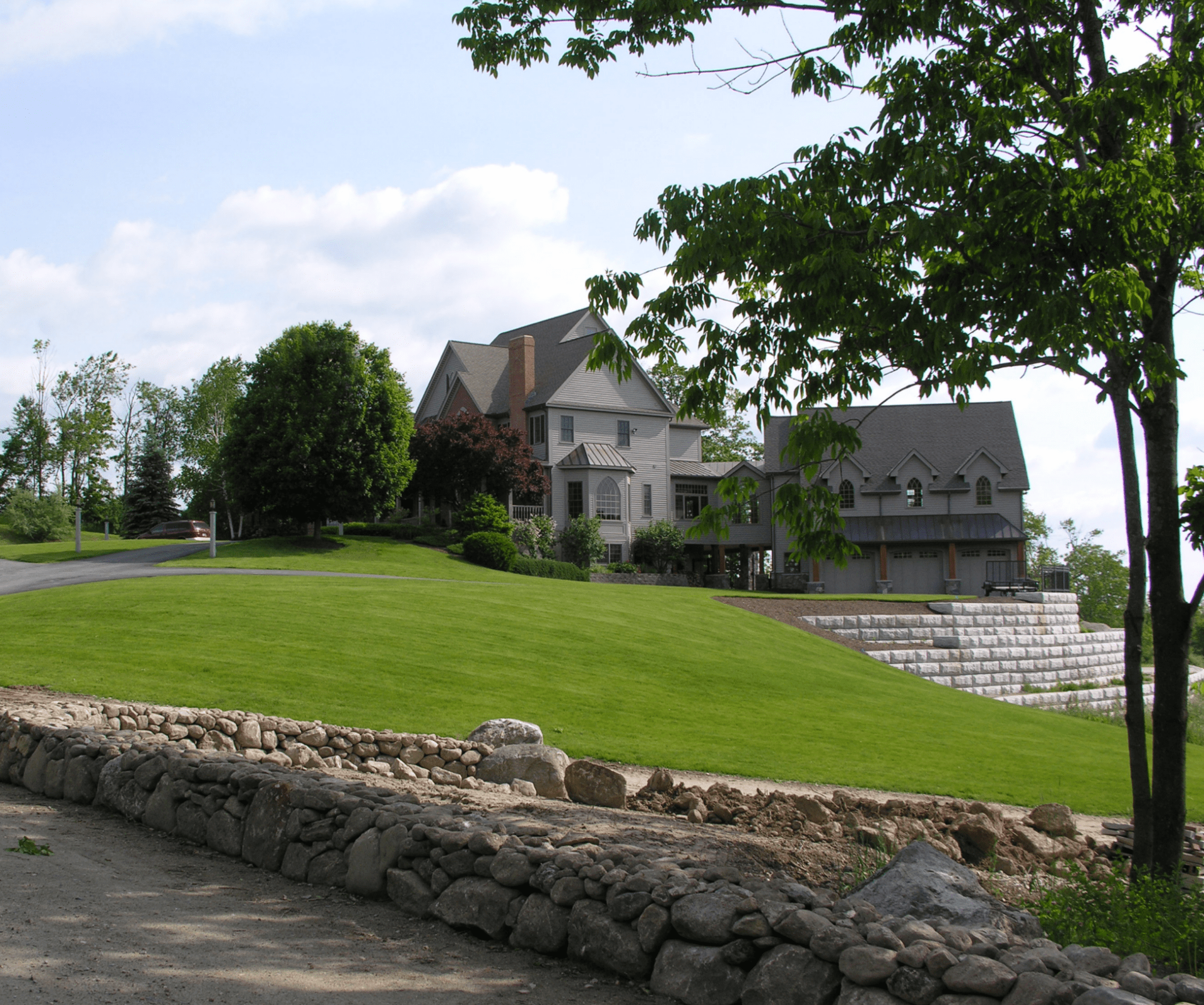 A large house sits on a lush green hill, with a stone wall in the foreground and trees surrounding the area.