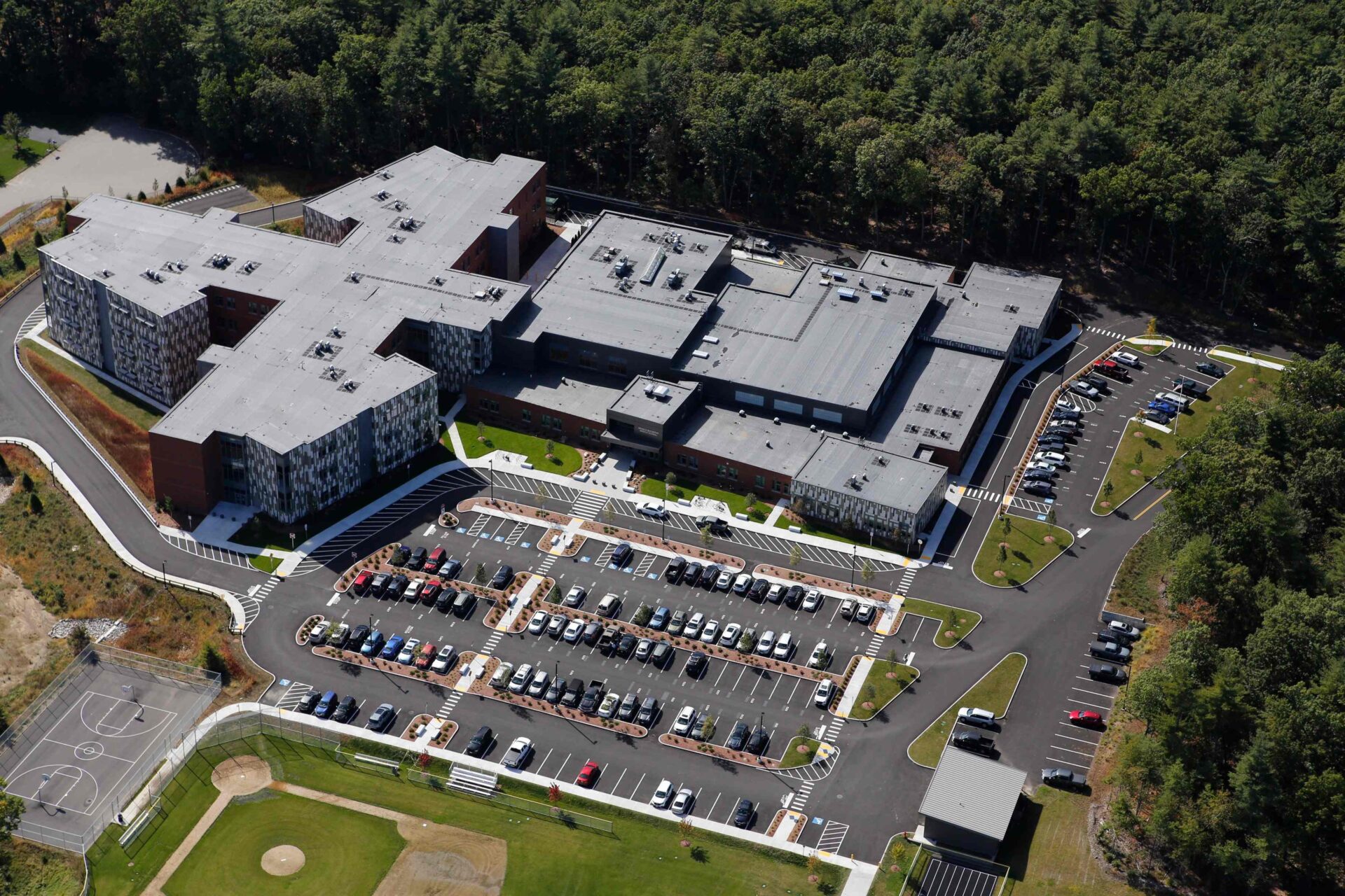 Aerial view of a large building complex surrounded by parking lots, forest, sports courts, and fields under a clear sky.