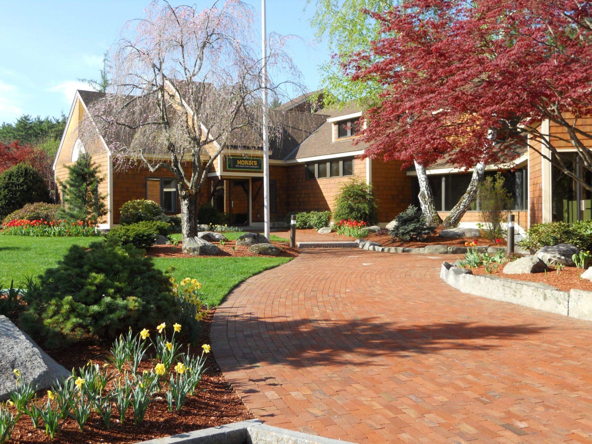 A charming building with a brick walkway is surrounded by lush gardens and blooming flowers under a sunny sky, creating a peaceful atmosphere.