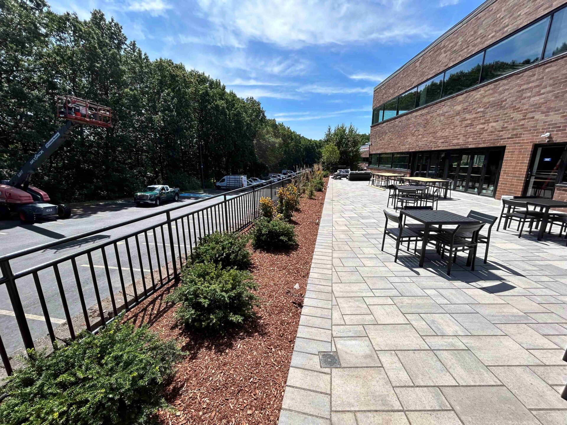 The image shows a stone patio with tables, a brick building, trees, and parked cars under a clear blue sky.