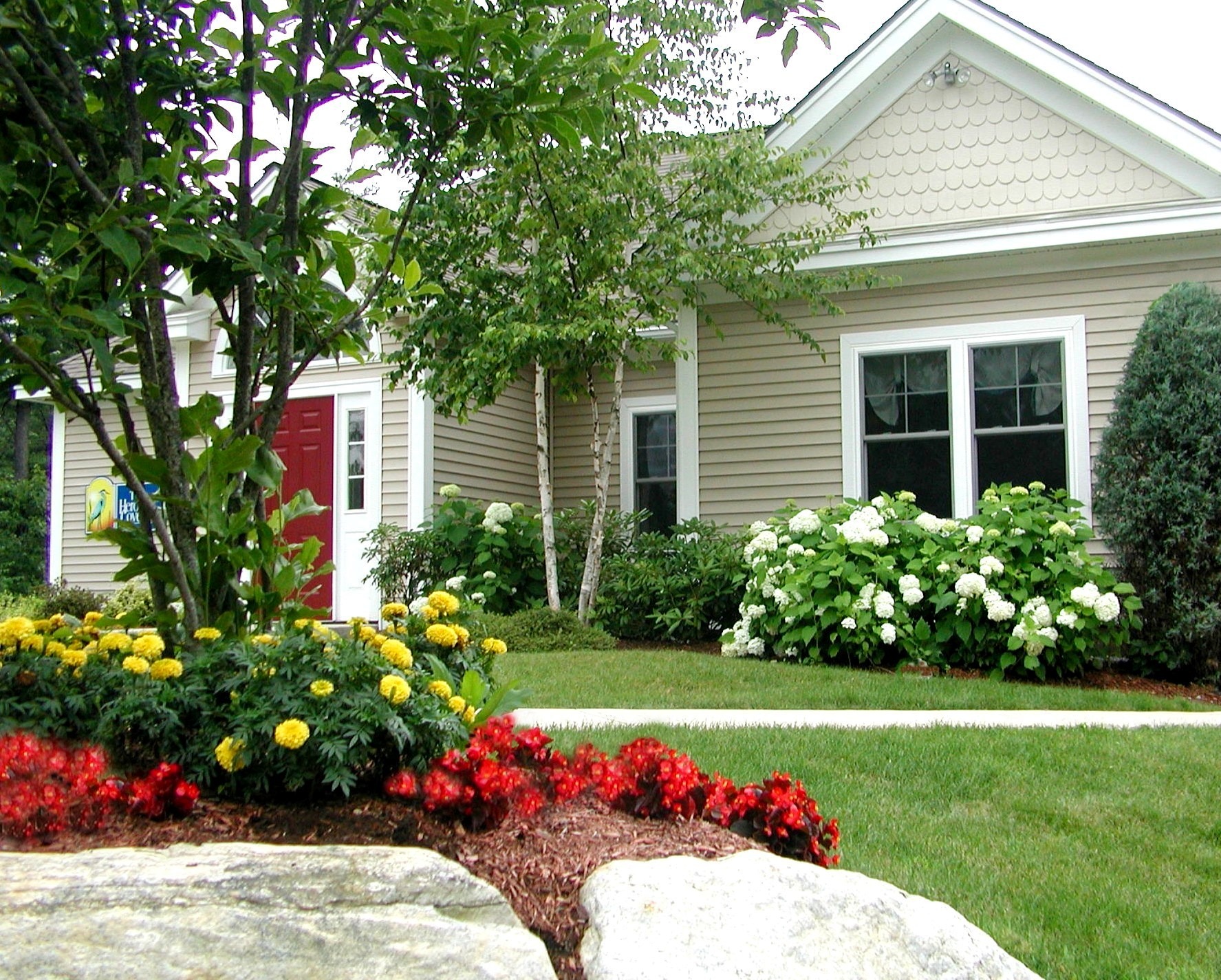 A charming house with light siding, surrounded by lush shrubs, vibrant flowers, and a well-maintained lawn. Red door adds a striking contrast.