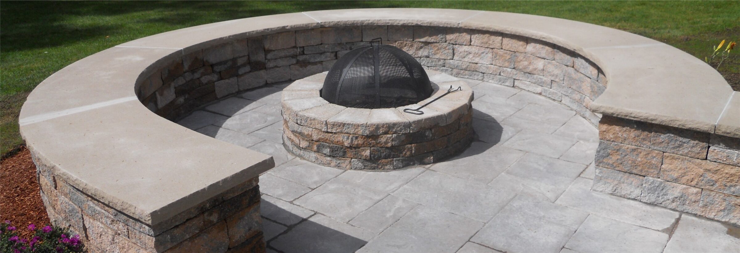A stone fire pit with a metal grate is surrounded by a curved stone bench, set on a paved patio with grassy surroundings.