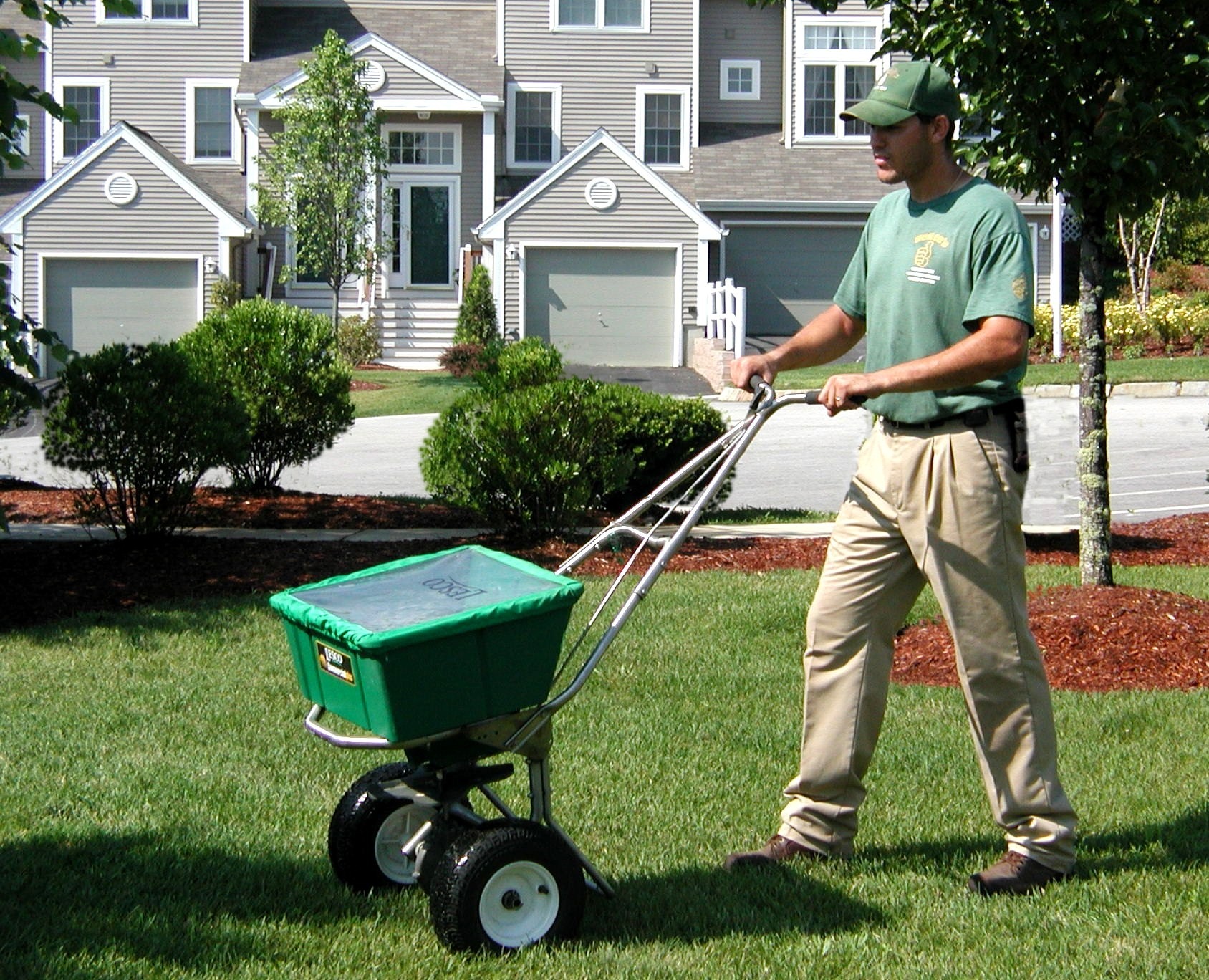 A person pushing a fertilizer spreader on a well-maintained lawn in front of a residential complex with neatly trimmed bushes.