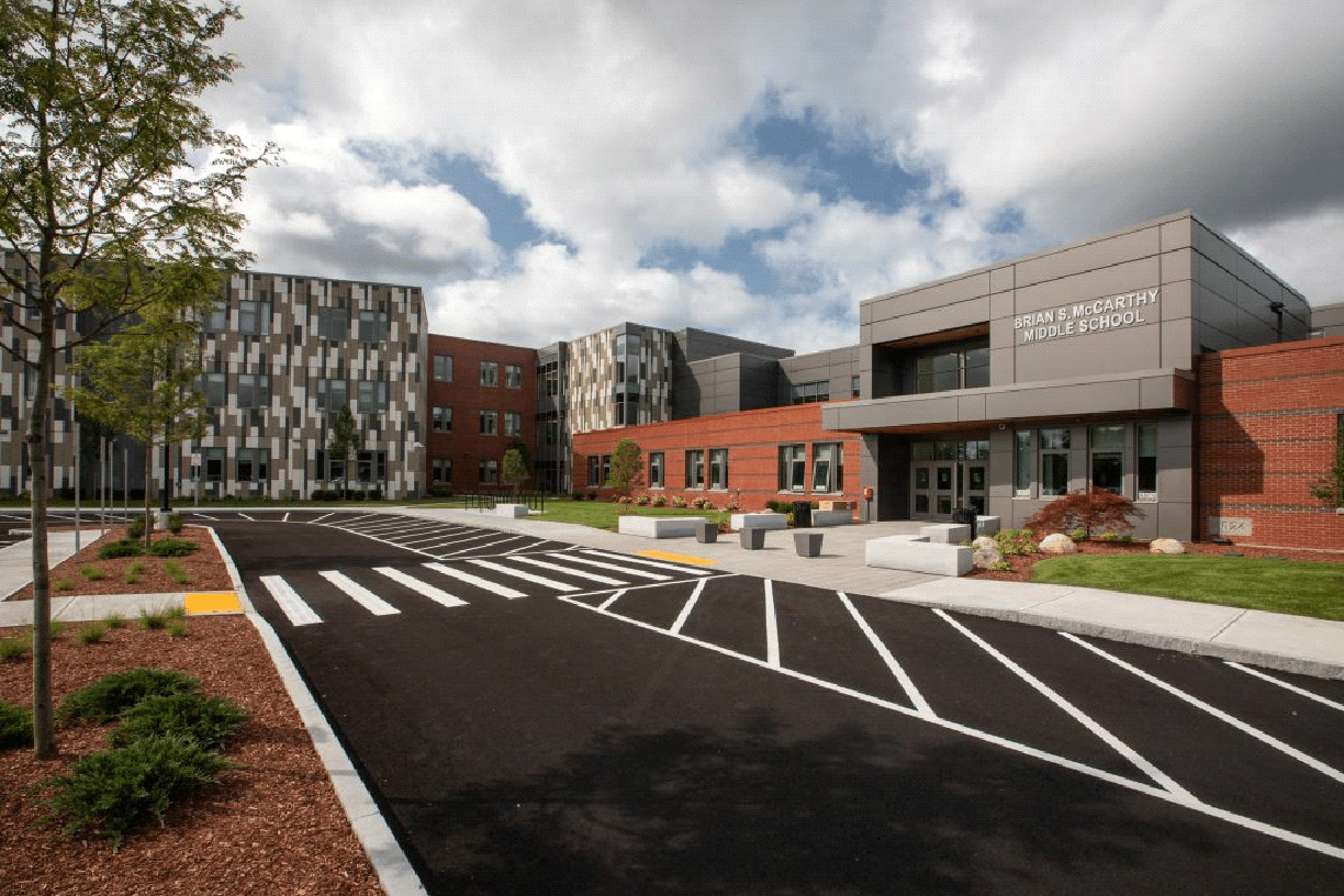 Modern school building with patterned facade, spacious parking area, and landscaped surroundings under a partly cloudy sky. No people visible.